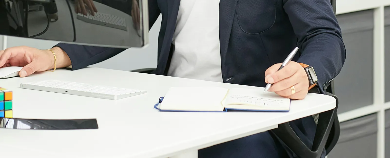 Man in glasses and blazer working at desk with computer, writing in notebook in a modern office setting.