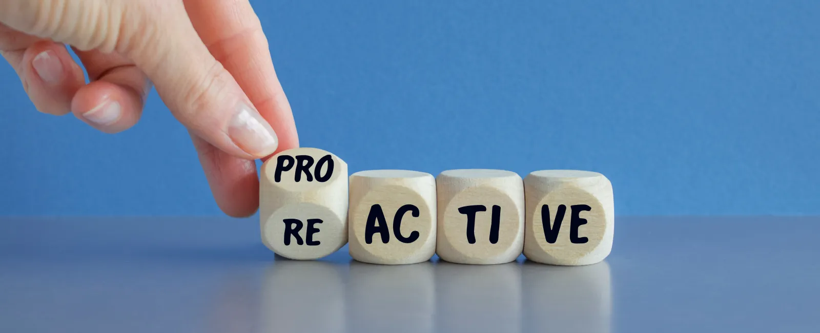 Hand turning wooden cube from RE to PRO to form proactive on blue background with reflective surface.