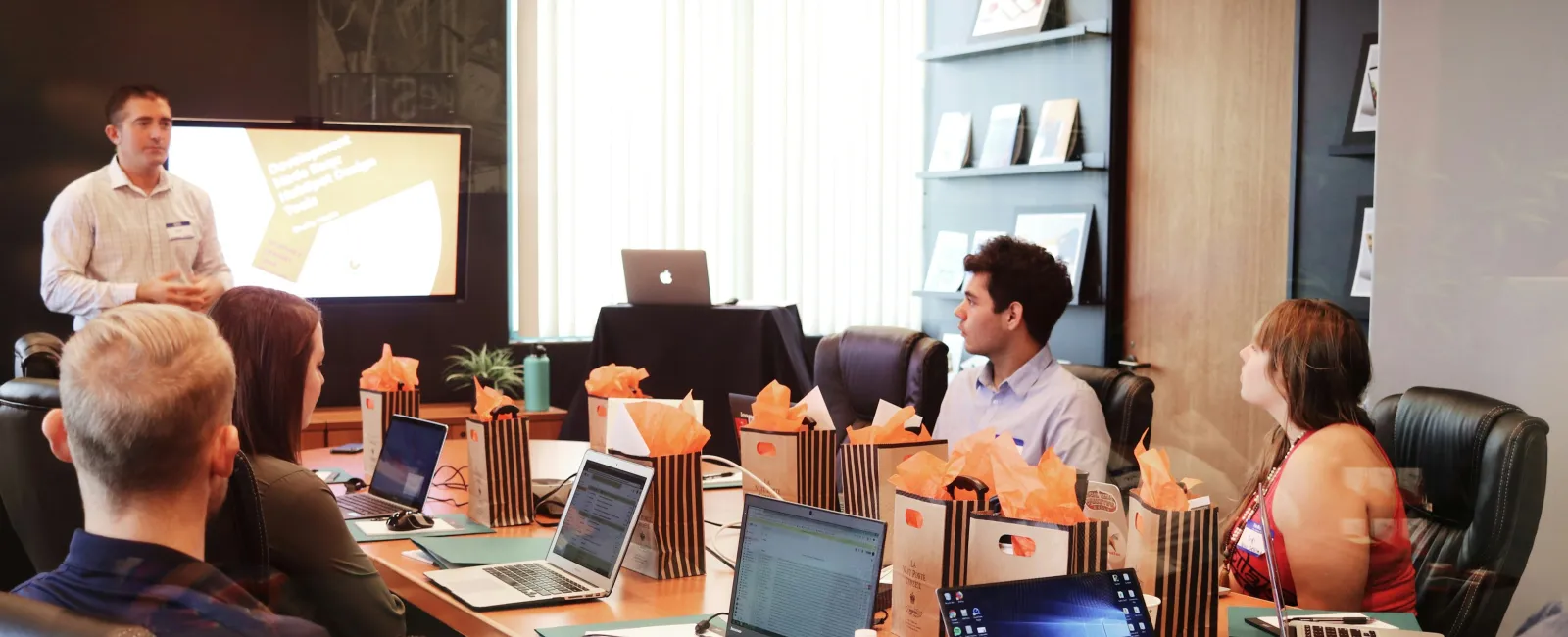 Team meeting in modern conference room with laptops, gift bags, and presenter using large screen display.