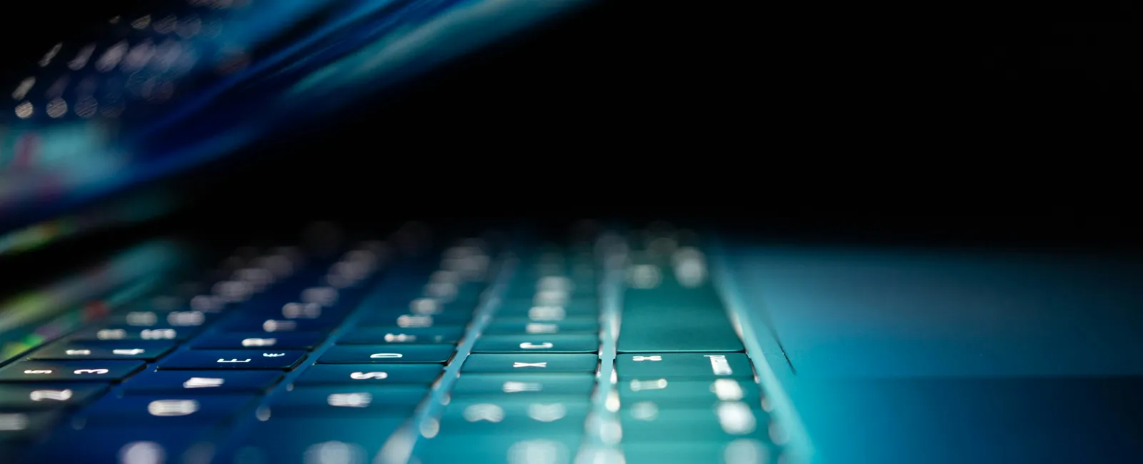 Close-up of a laptop keyboard with glowing backlit keys reflecting on the screen in low light.
