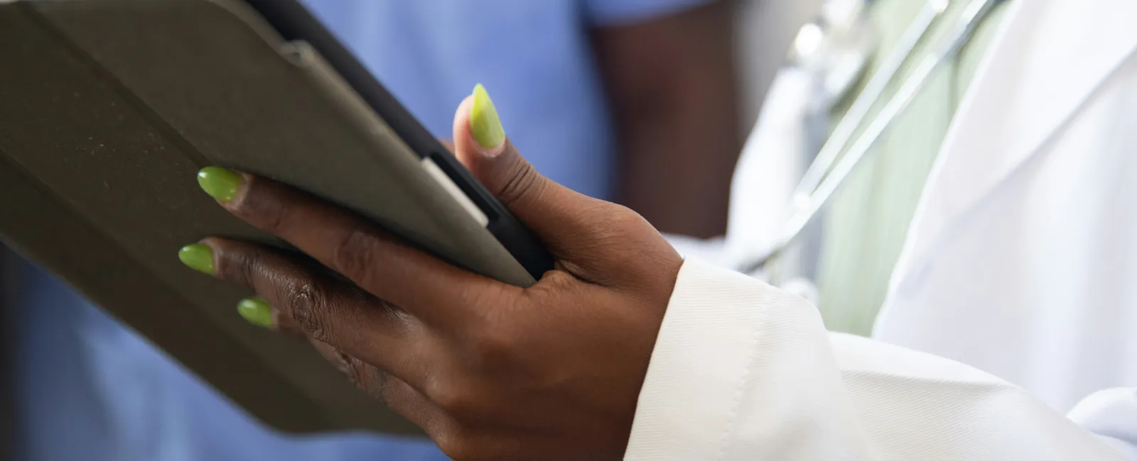 Close-up of a doctor with green nails using a tablet, wearing a white coat and stethoscope.