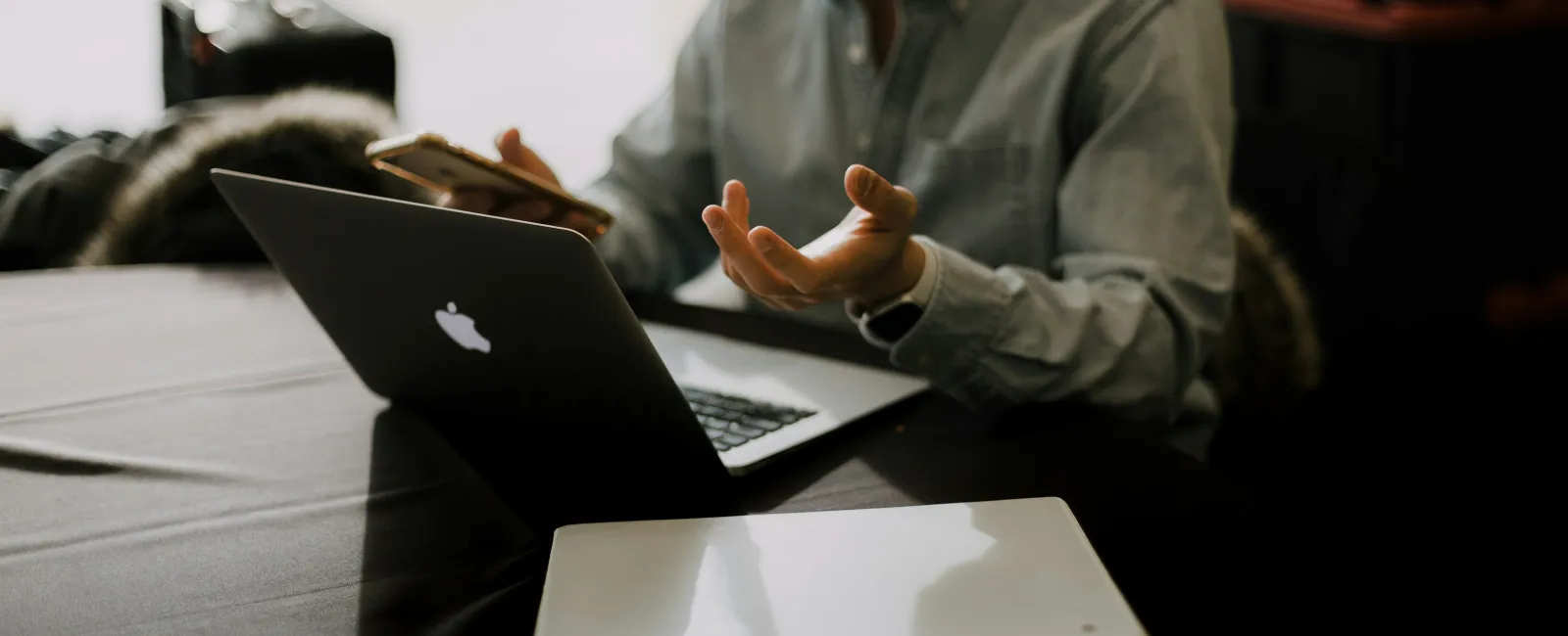 Person using a MacBook laptop and smartphone while discussing or explaining at a table with a white box.