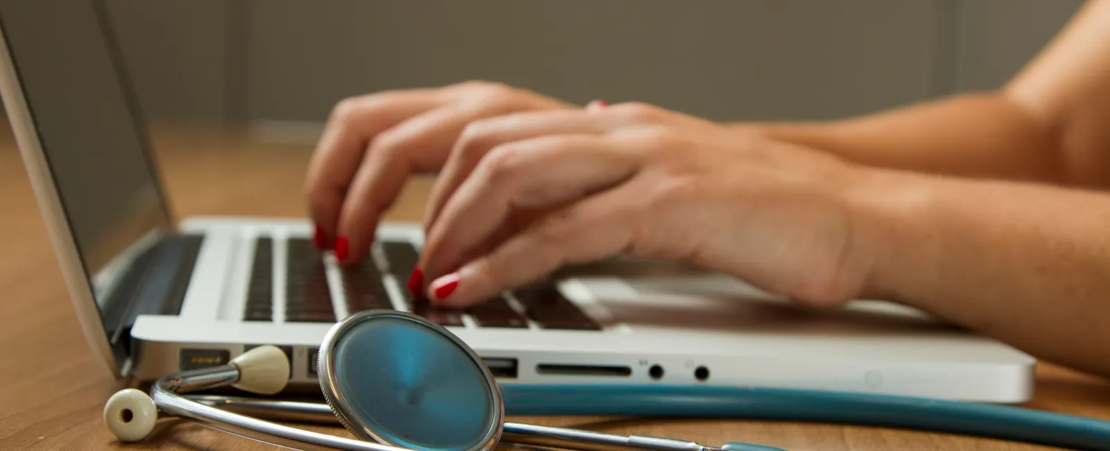 Hands typing on a laptop keyboard with a stethoscope on the wooden desk, symbolizing healthcare and technology.