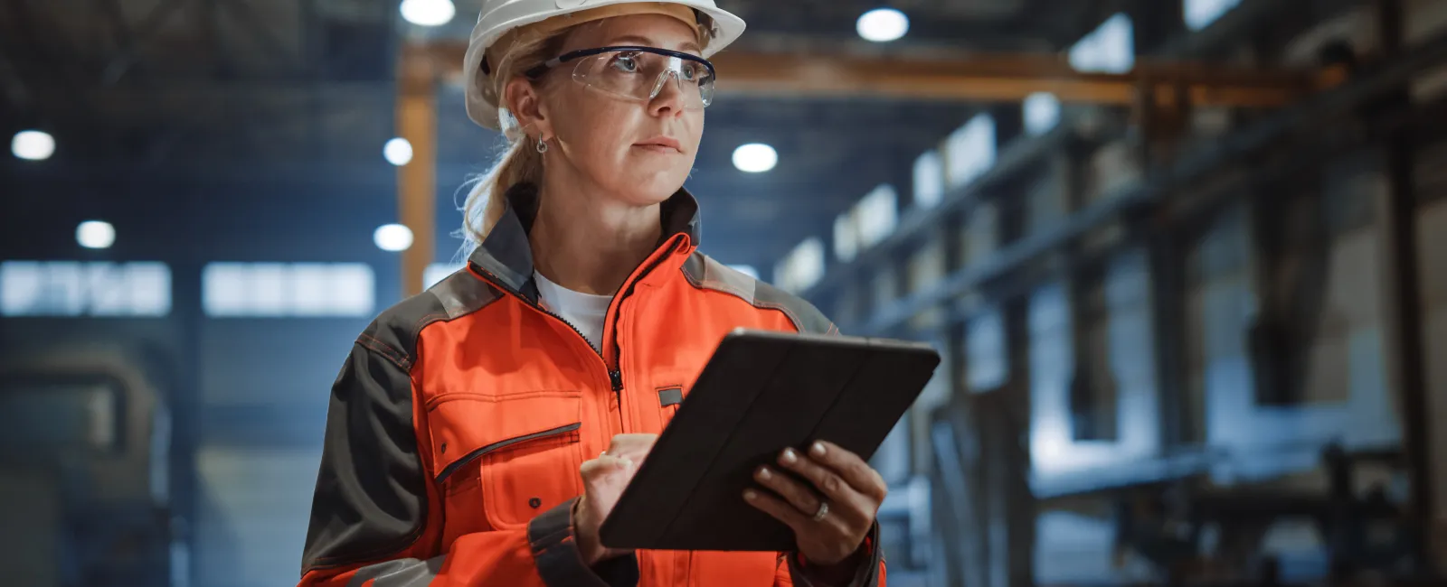Female engineer in safety gear using tablet inside industrial factory with machinery and equipment in background.