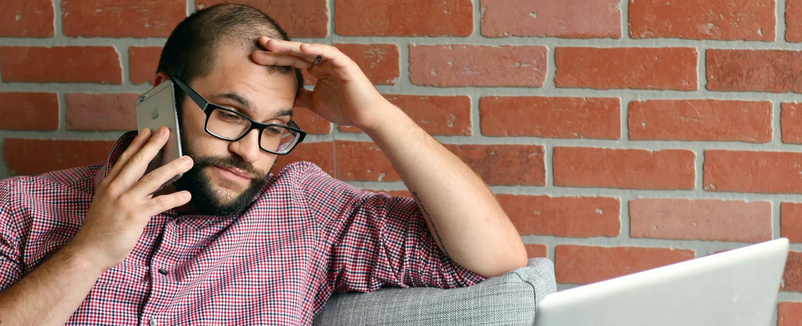 Man in glasses sitting on couch talking on phone with worried expression and laptop open against brick wall