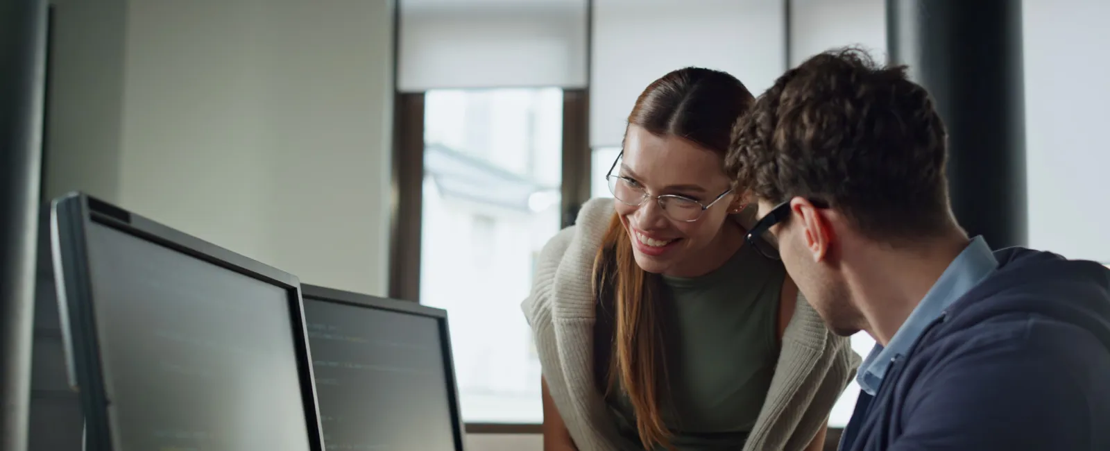 Two colleagues collaborate at a desk, reviewing code on dual computer monitors in a modern office.