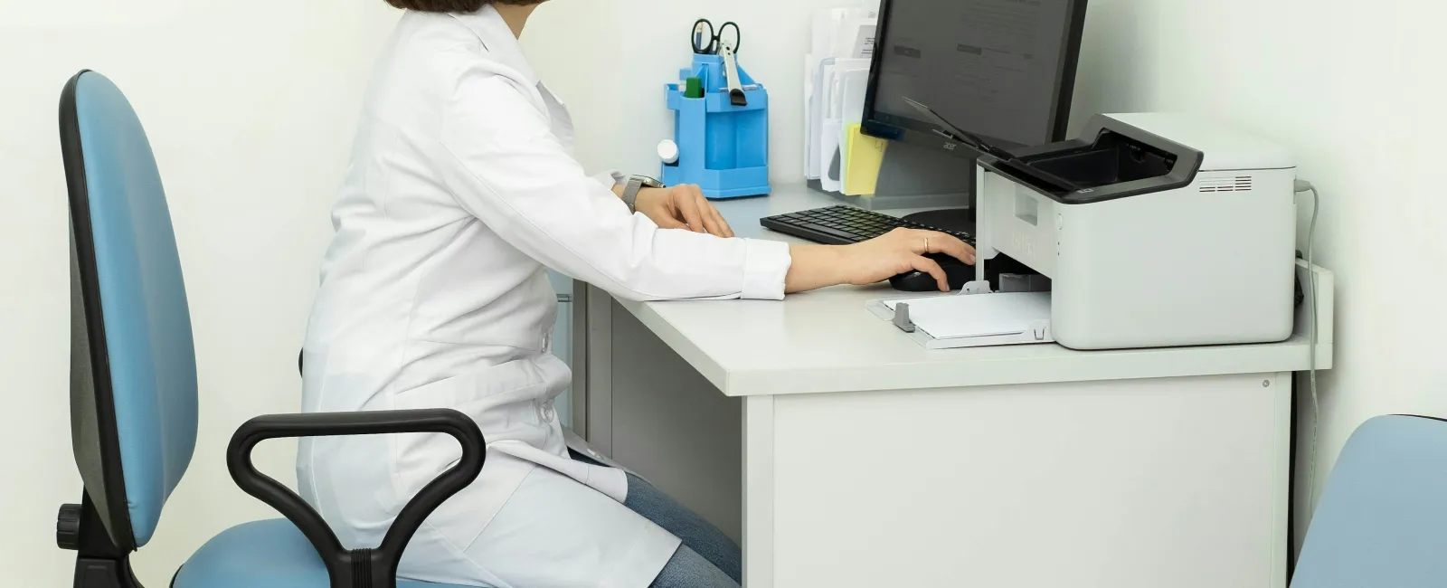 Female doctor in white coat working on computer at a clean, modern medical office desk.