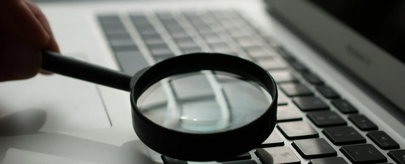 Magnifying glass held over laptop keyboard, highlighting keys in soft light and shadow.