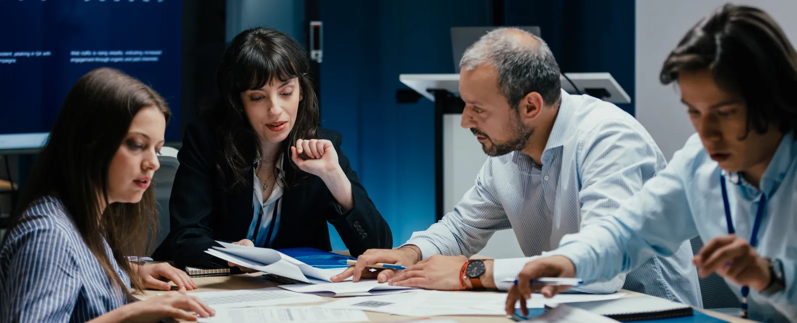Four professionals review documents during a business meeting with data charts displayed on screen in a modern office.