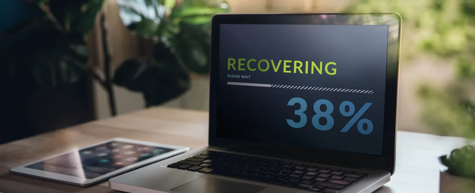 Laptop on desk showing data recovery progress at 38% with tablet beside it and plants in background.