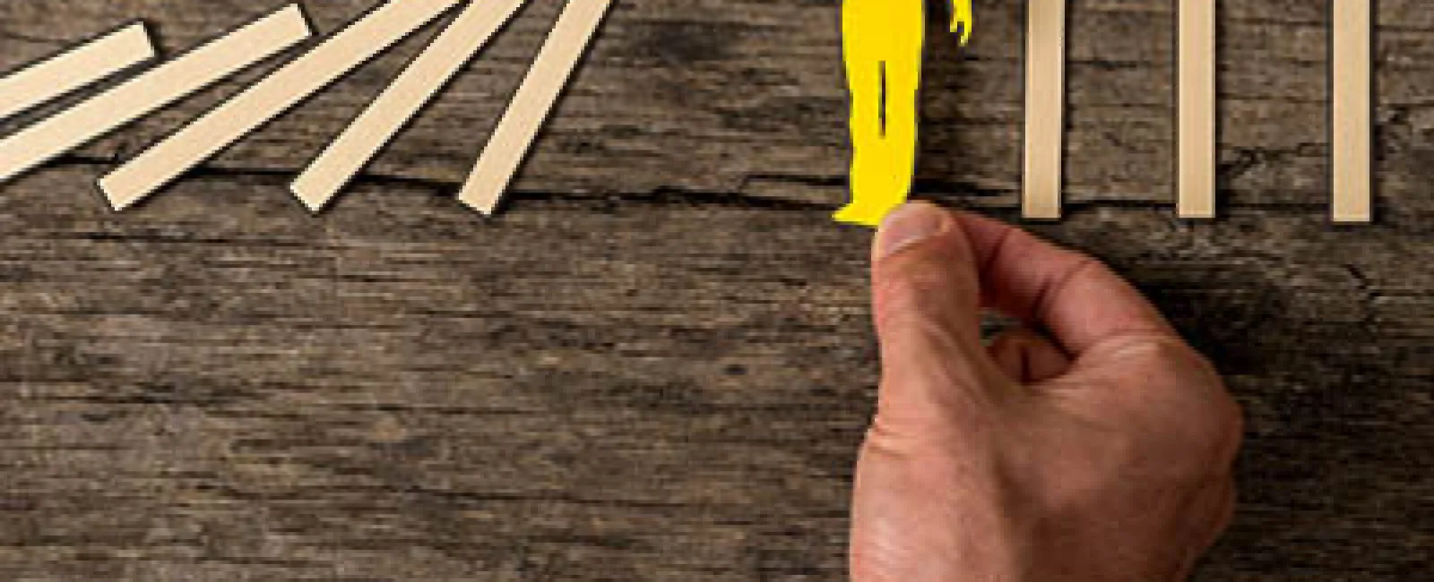 Hand holding a yellow figure stopping falling dominoes on a wooden surface symbolizing prevention or intervention.