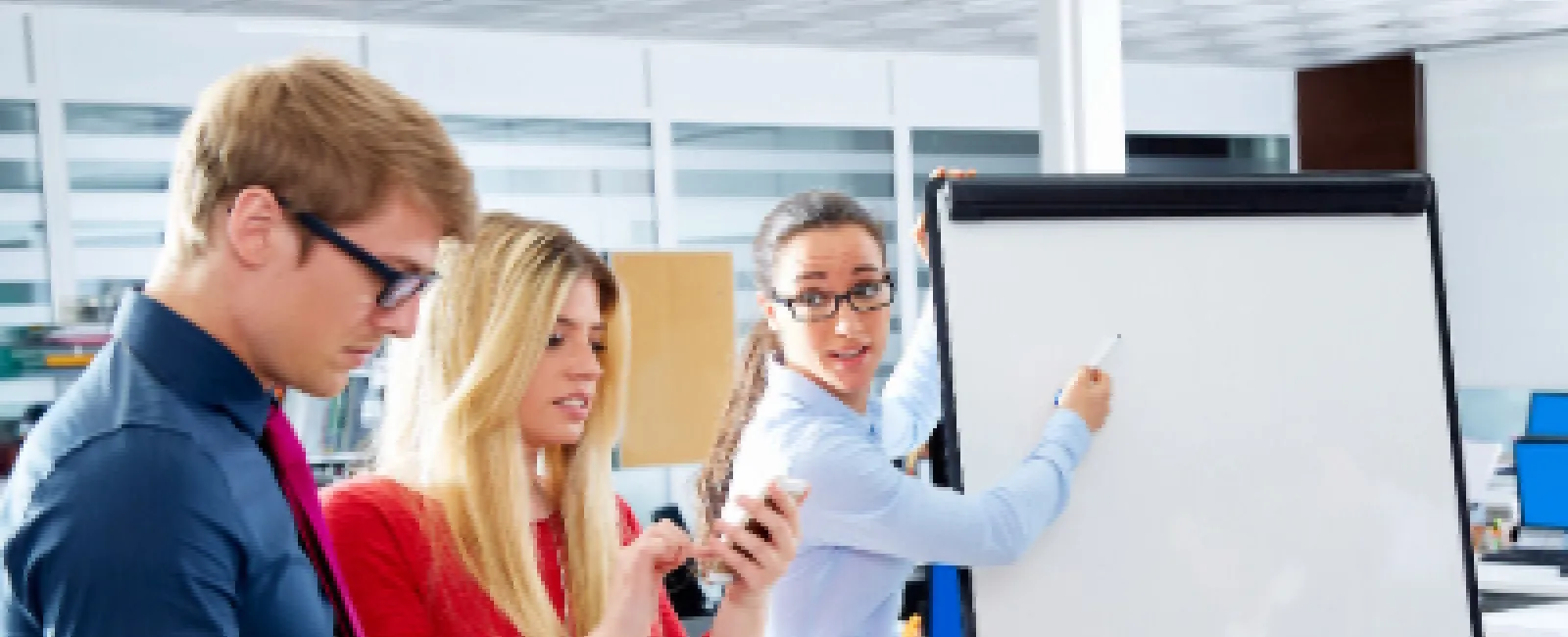 Three professionals in office discussing and using digital devices with a whiteboard chart in a modern workspace.