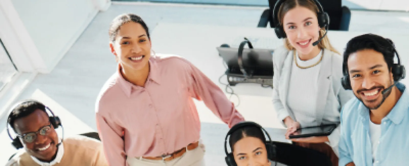 Diverse customer support team wearing headsets smiling in a modern office with laptops and notebooks.