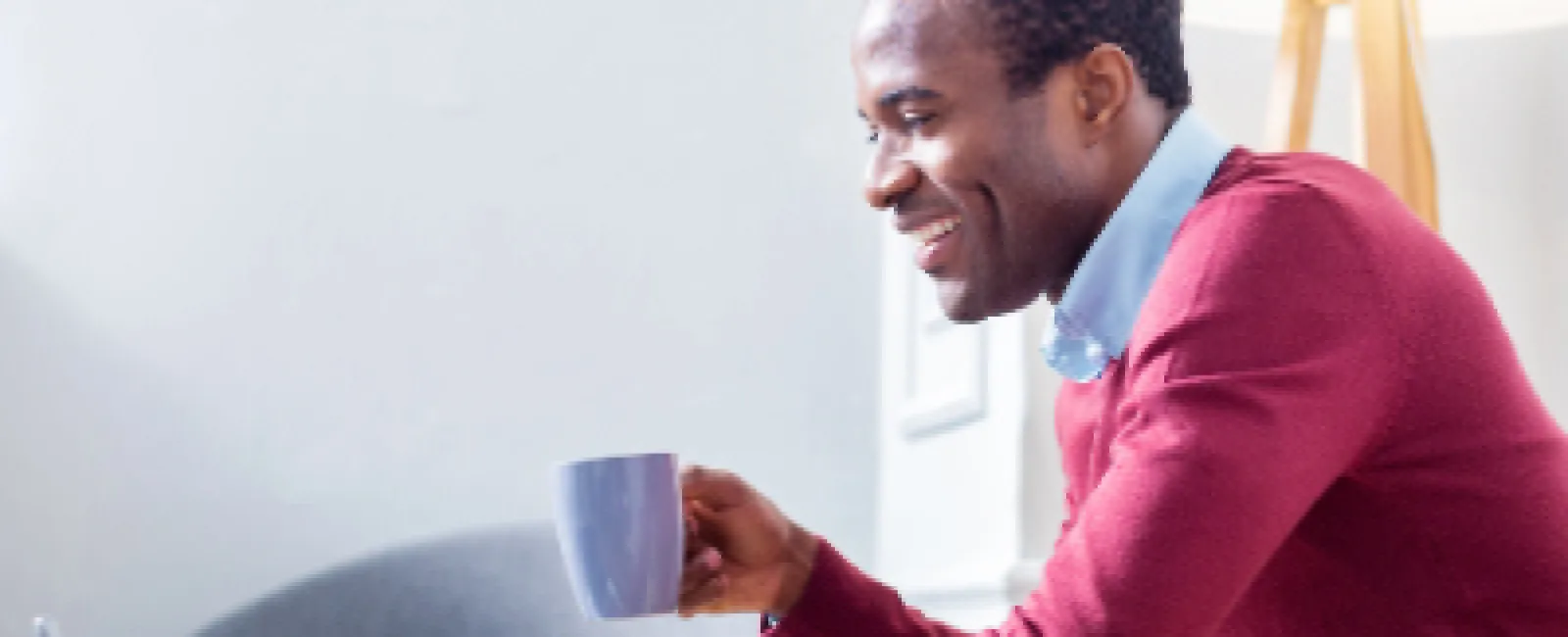 Man in red sweater sitting on couch, smiling while using laptop and holding a blue mug indoors.