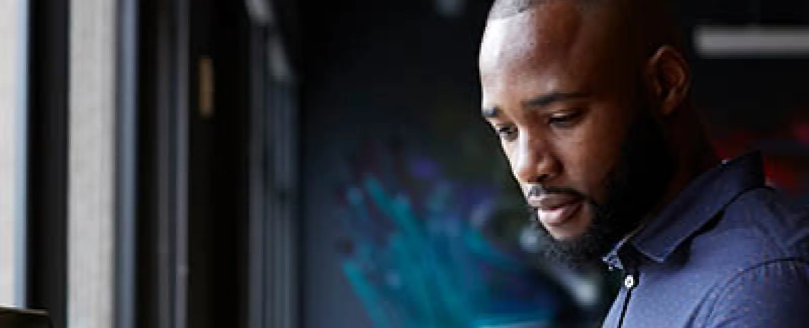 Man in blue shirt using smartphone and holding coffee cup while sitting at wooden desk with laptop.