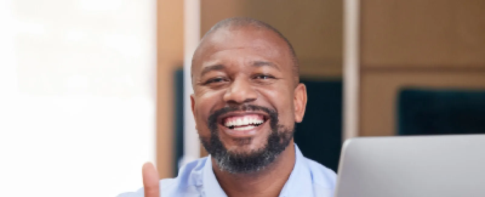 Smiling man in blue shirt giving thumbs up while sitting at desk with computer and smartphone nearby
