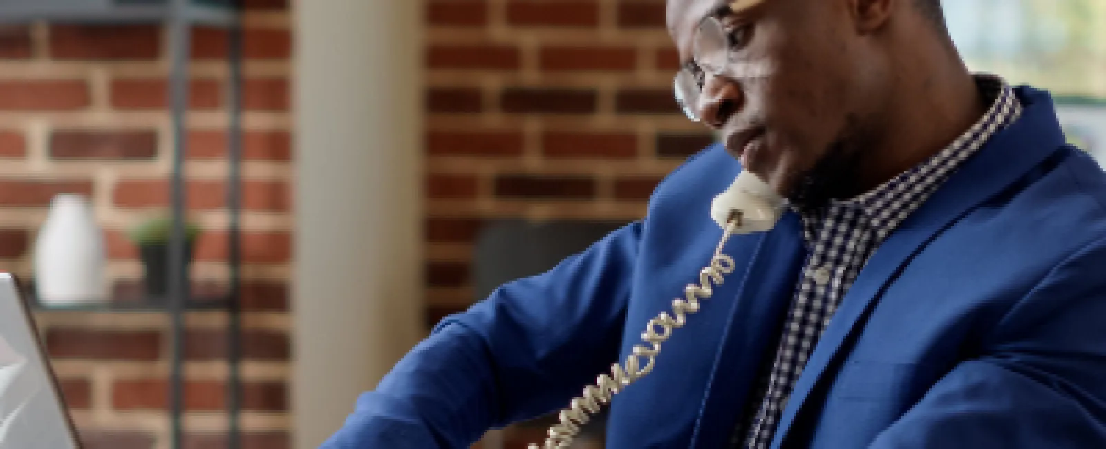 Professional man in blue suit multitasking with phone and laptop in modern office space with brick wall.