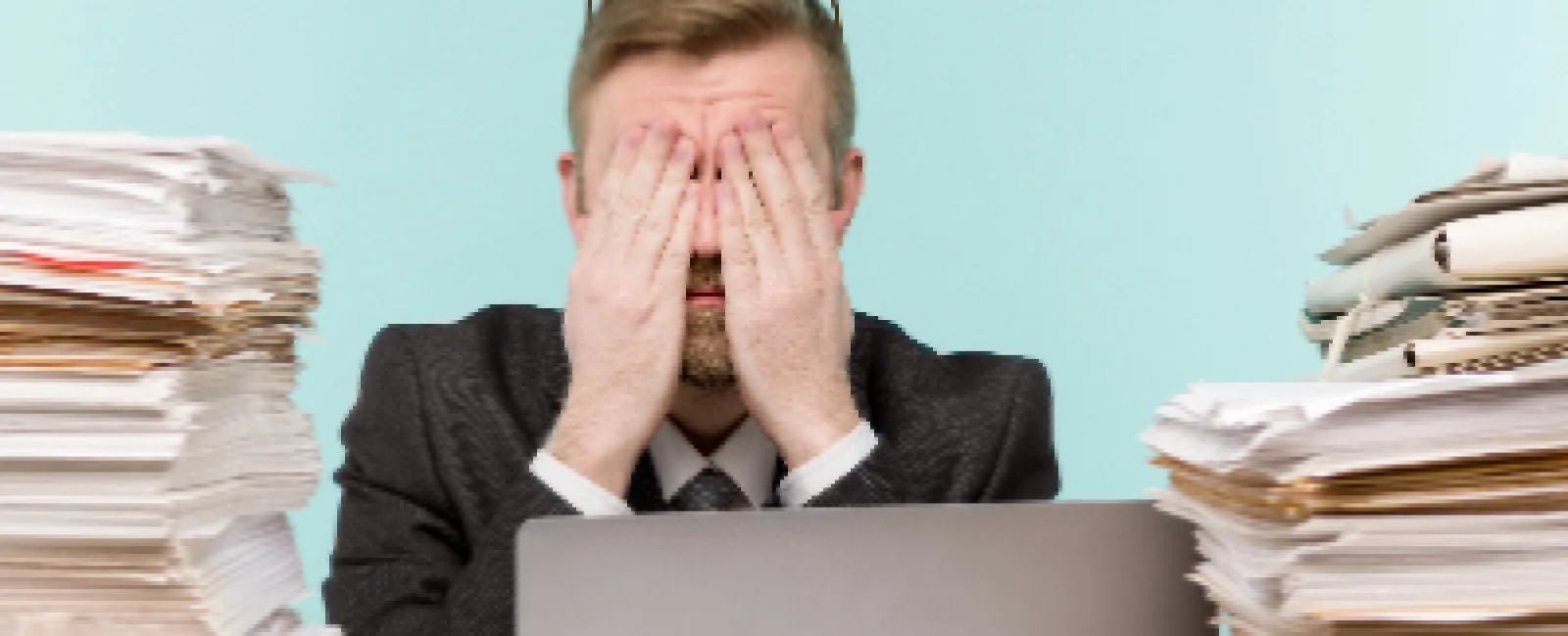 Stressed businessman covering his face sitting between tall stacks of paperwork with a laptop on desk.