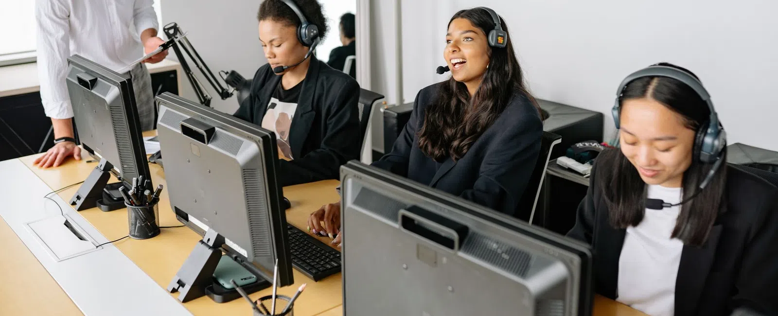Diverse call center agents with headsets working at computers and a supervisor assisting in a bright office.