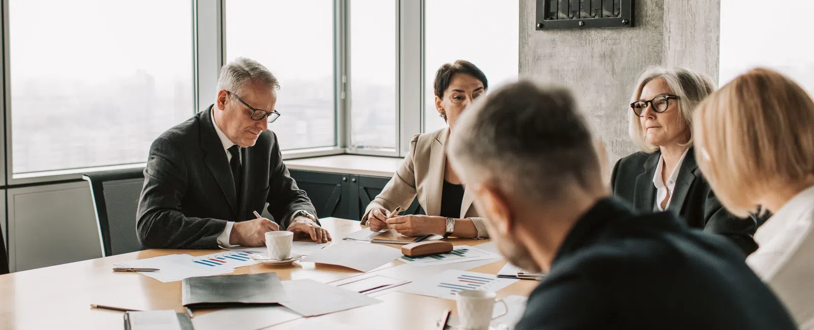 Business team in formal attire discussing documents and taking notes during office meeting by large windows.