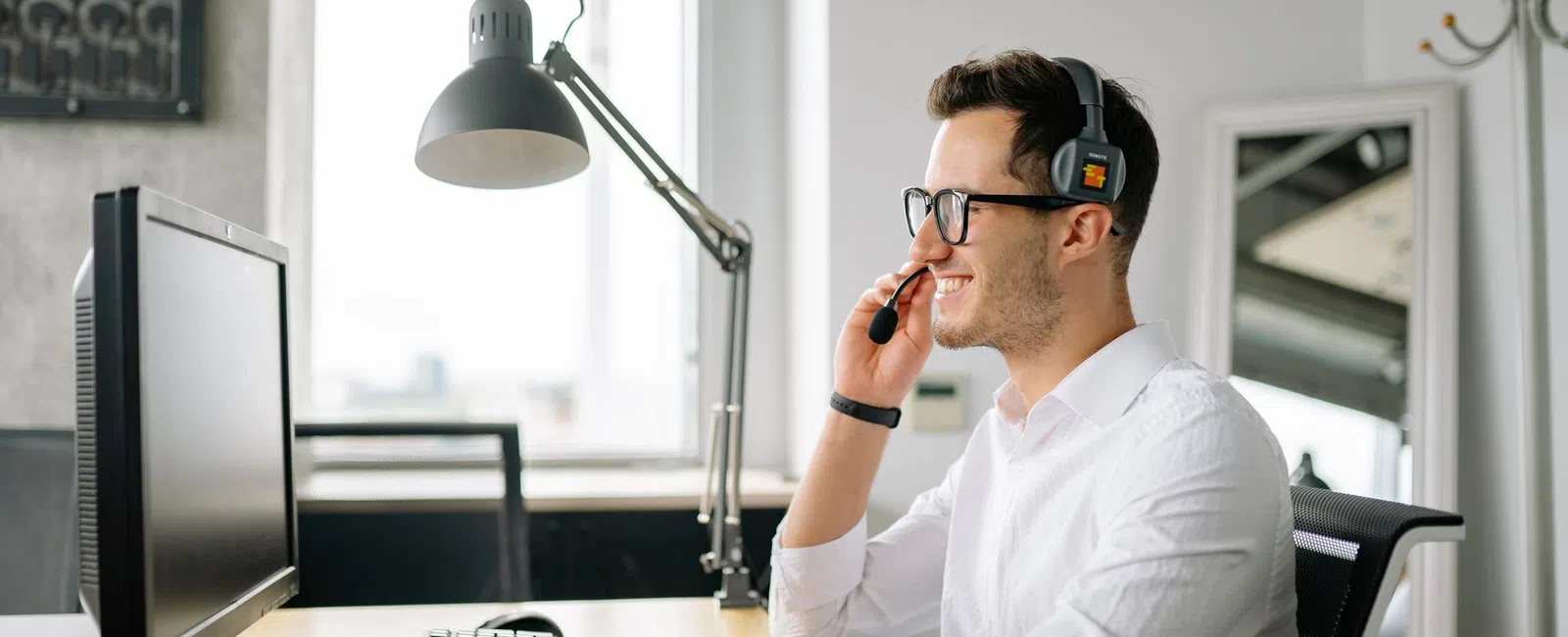 Smiling man in white shirt wearing headset working on computer in bright modern office workspace