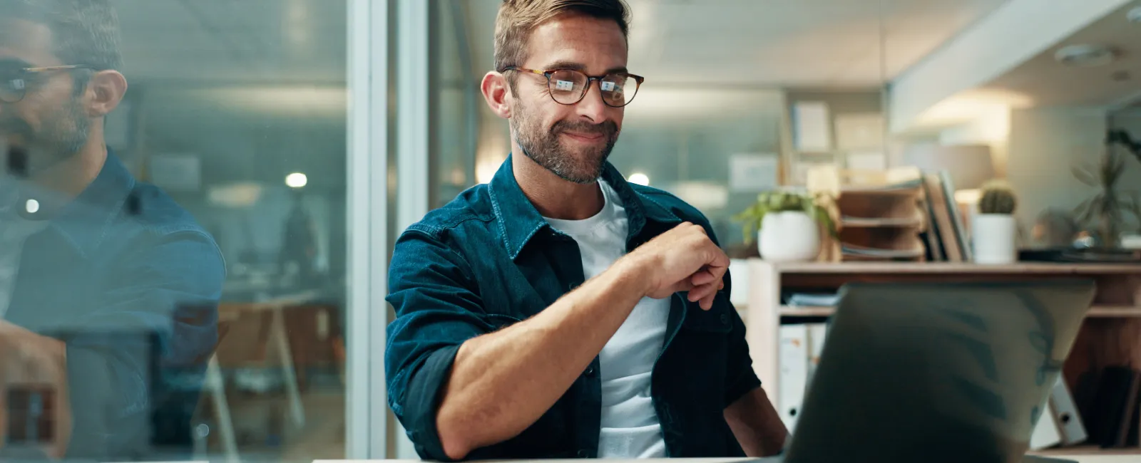 Smiling man wearing glasses working on a laptop inside a modern office with tablet on desk.