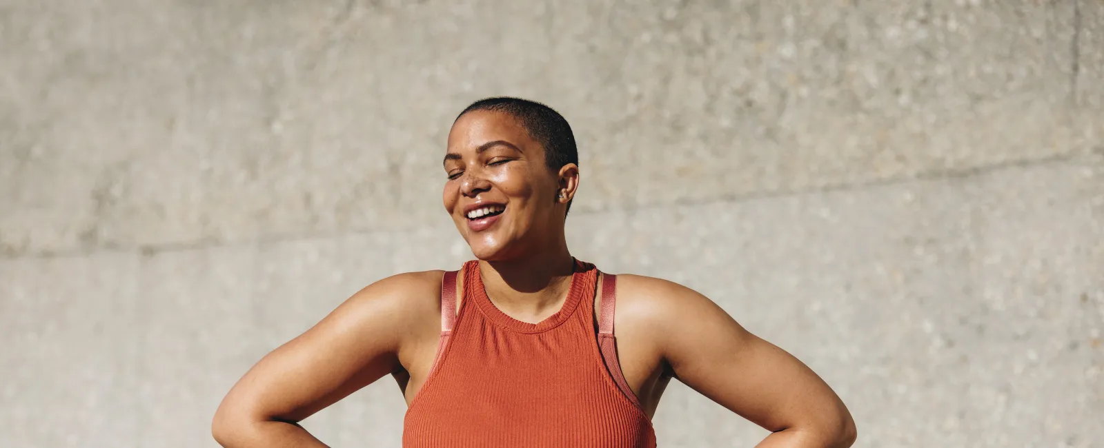 Smiling woman in burnt orange workout clothes poses confidently against a neutral concrete wall background.