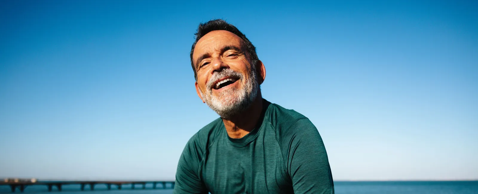 Happy elderly man in green shirt resting by the water with a fitness bottle under clear blue sky.