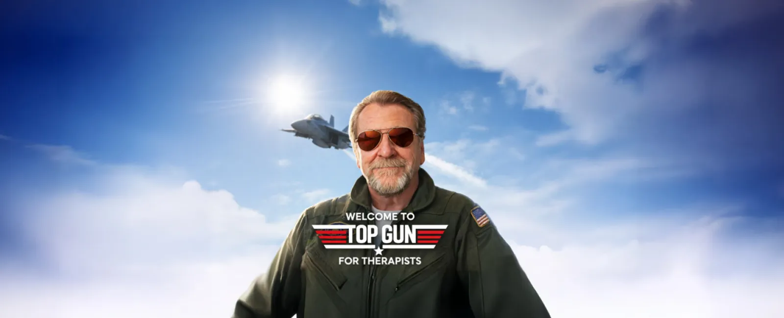 Man in pilot suit with sunglasses stands under a bright sky with a fighter jet flying behind him