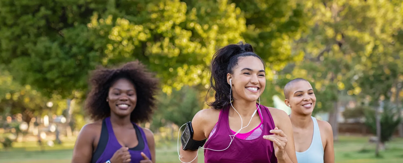 Three women jogging outdoors in athletic wear, smiling and enjoying a sunny day in a green park.
