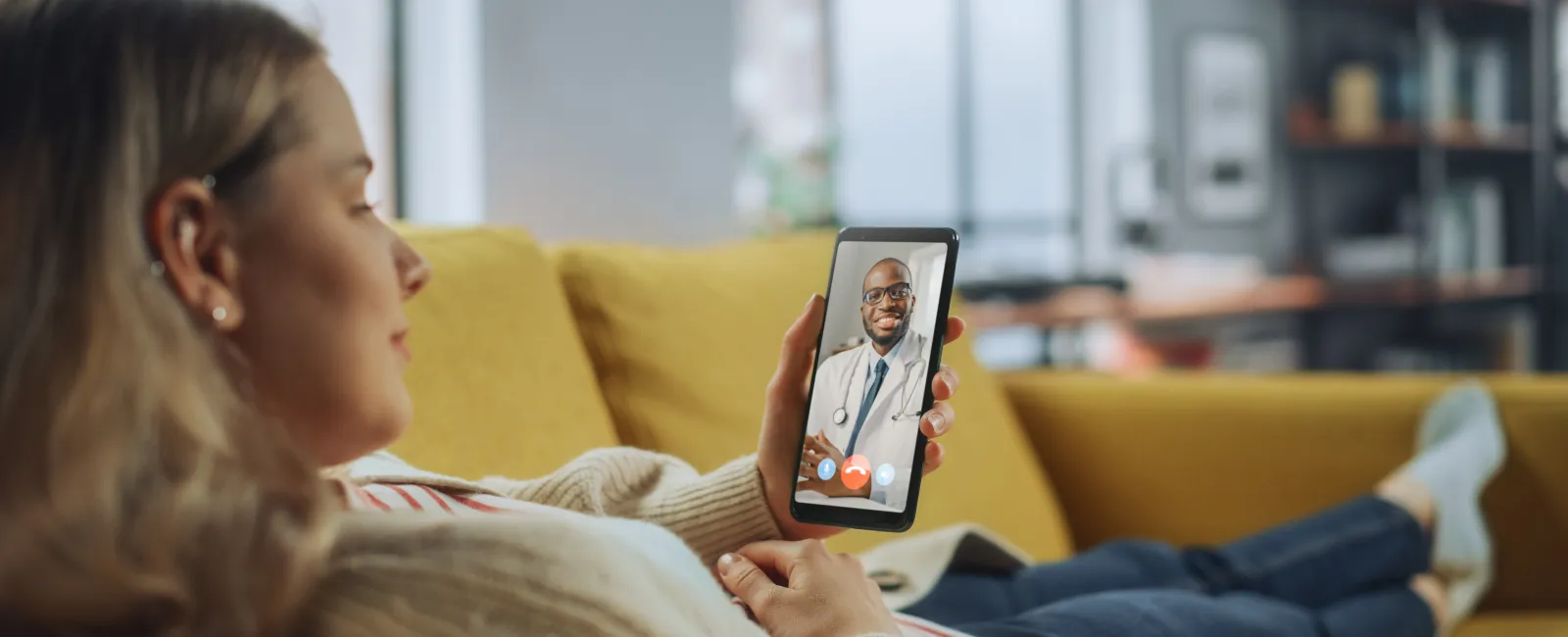 Woman having a video call with her primary care doctor on a smartphone while lying on a yellow couch at home