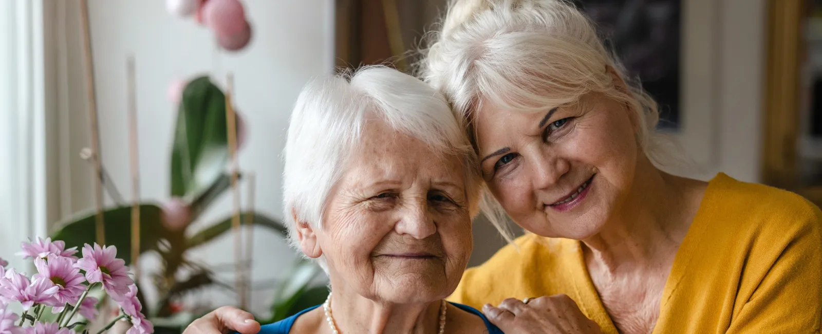 A warm, joyful moment between two older women smiling together surrounded by flowers.