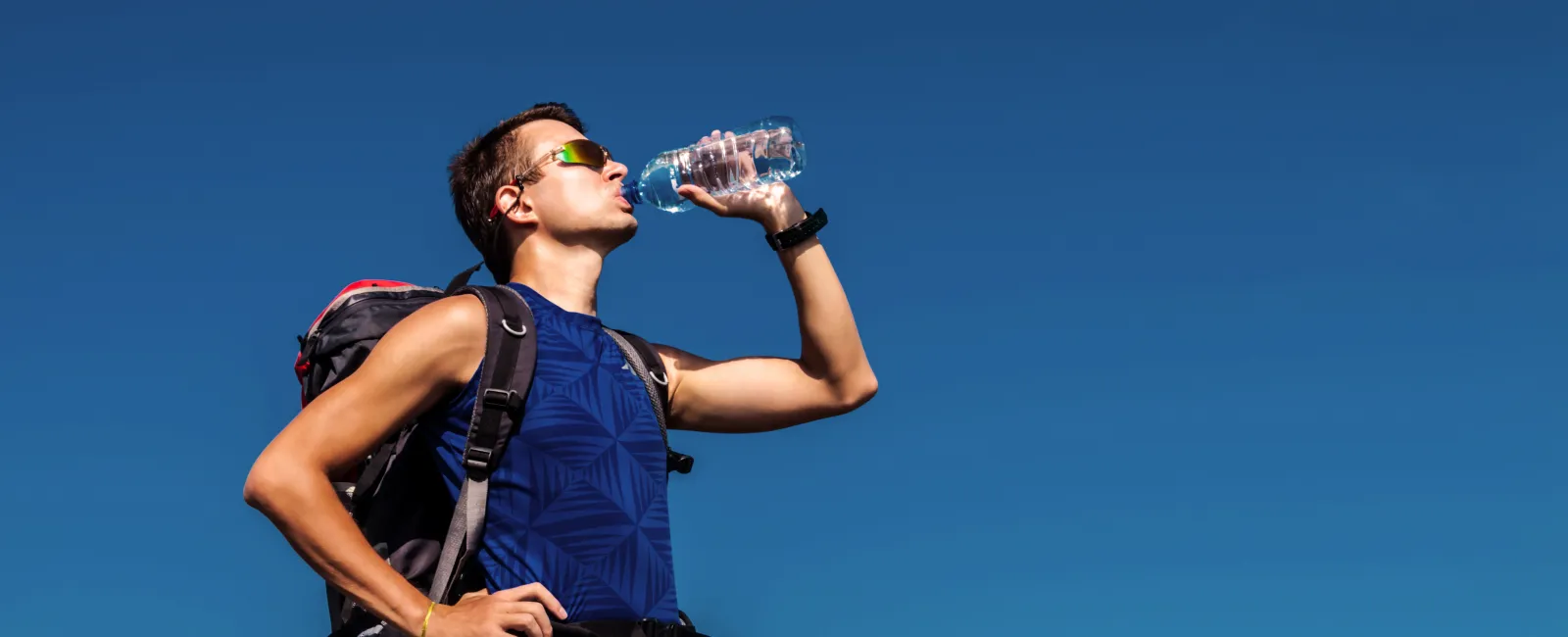 Hiker in sunglasses drinks water from a bottle against a clear blue sky during outdoor adventure.