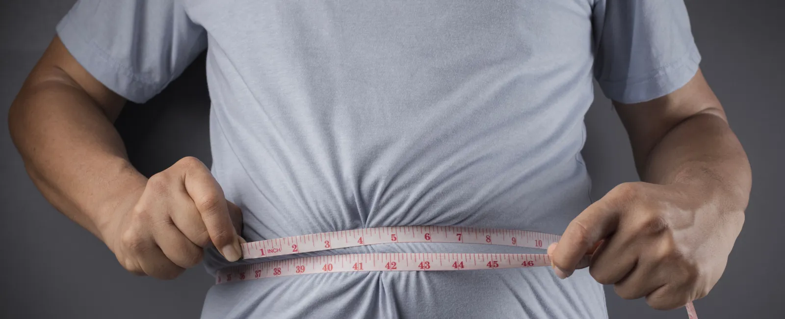 Person measuring waist with a pink tape measure over a gray shirt and blue jeans against dark background.