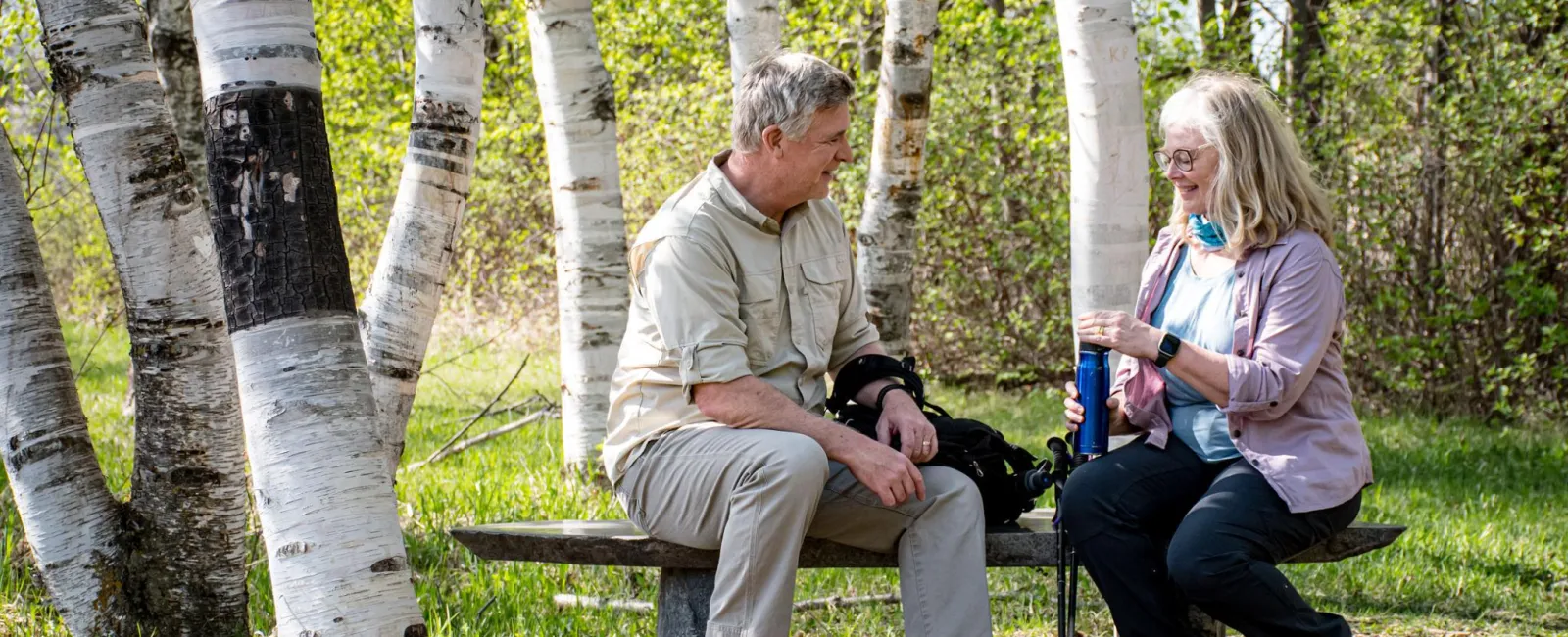 Older couple sitting on a wooden bench in a birch forest enjoying a break from hiking on a sunny day.
