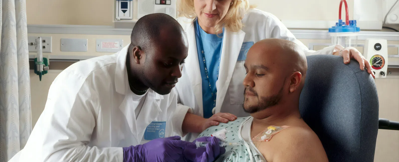 Two medical professionals attending to a seated male patient with an IV catheter in a clinical setting.