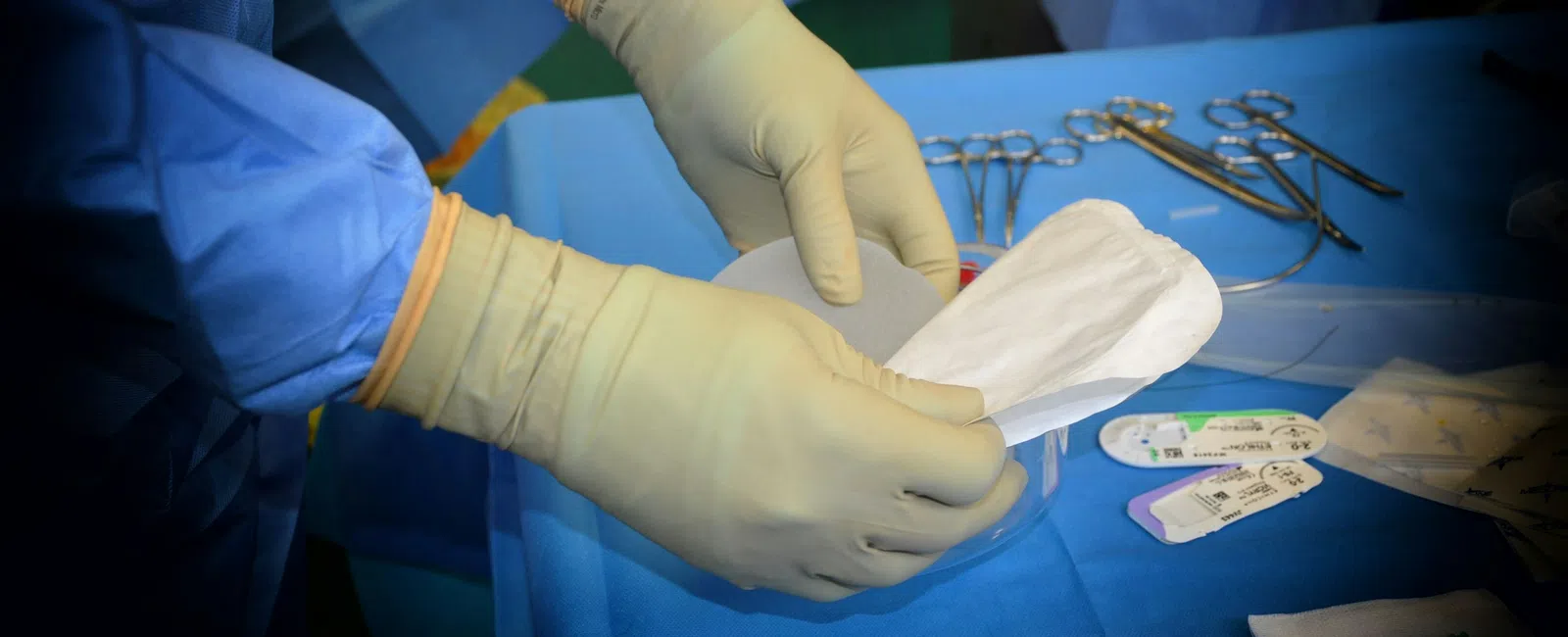 Surgeon wearing gloves handling a breast implant in a sterile operating room with surgical instruments nearby.