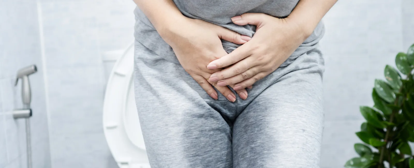 Person in gray clothes holding lower abdomen in pain sitting near a toilet in a bathroom.