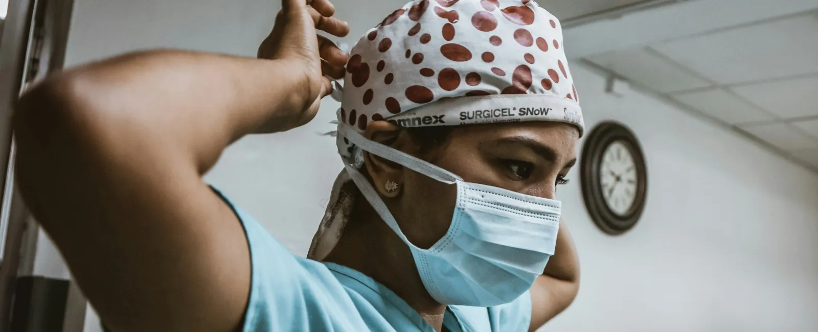 Healthcare worker in blue scrubs and polka dot surgical cap adjusting mask in hospital hallway.
