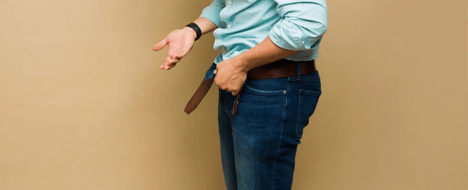 Man in blue checkered shirt and jeans adjusting his brown belt against beige background.