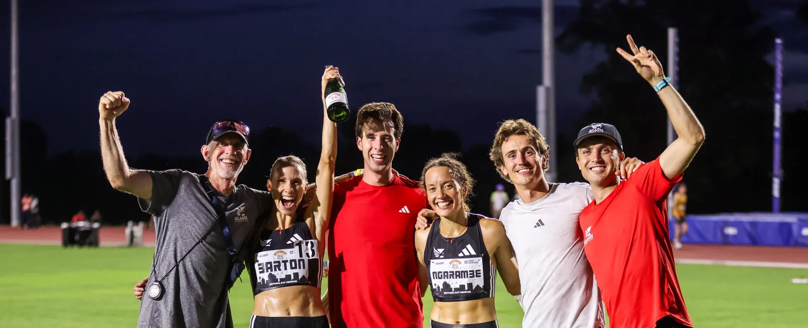 Group of six athletes celebrating on a track field at night, smiling and raising arms in victory.