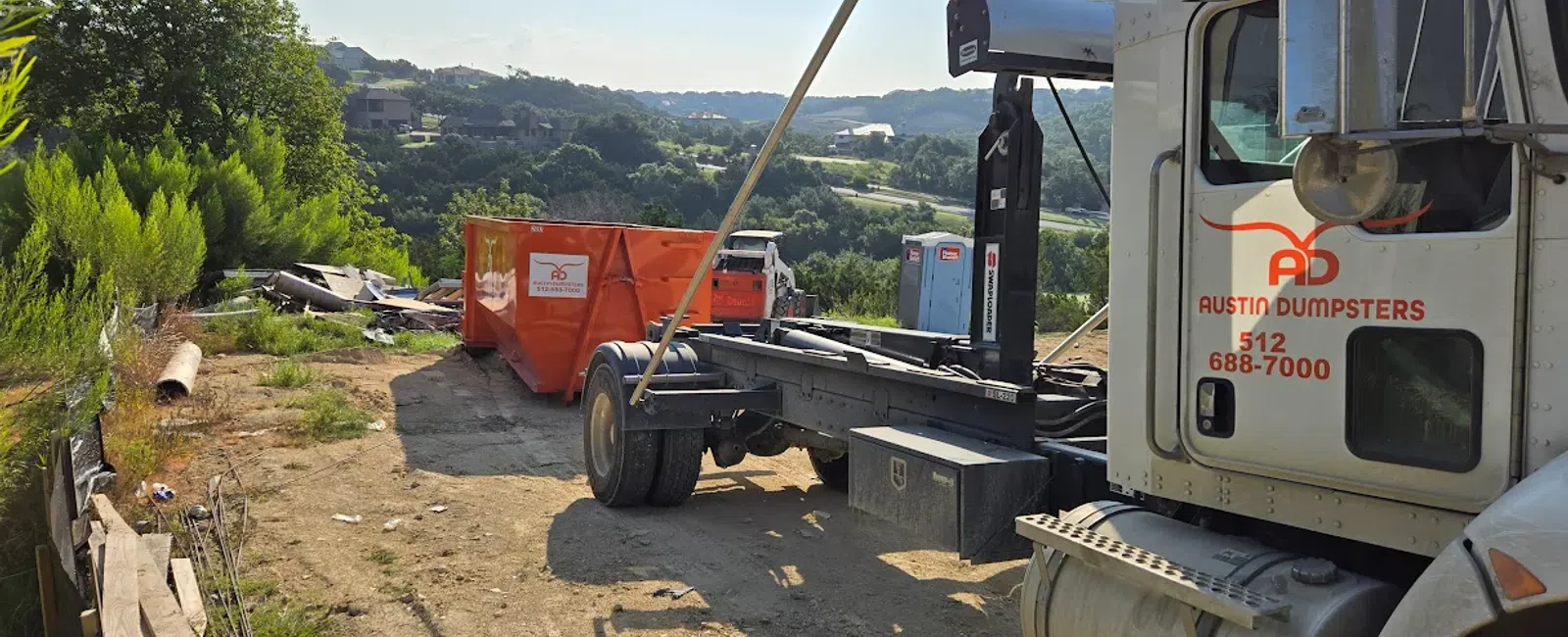 Austin Dumpsters truck unloading orange dumpster at a rural site with hills and greenery in the background.