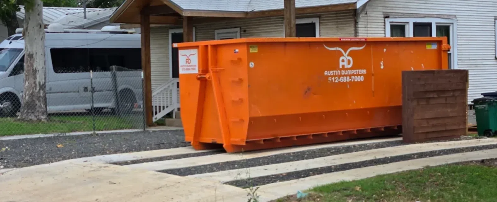 Large orange dumpster from Austin Dumpsters placed on a concrete pad in front of a white house with trees.