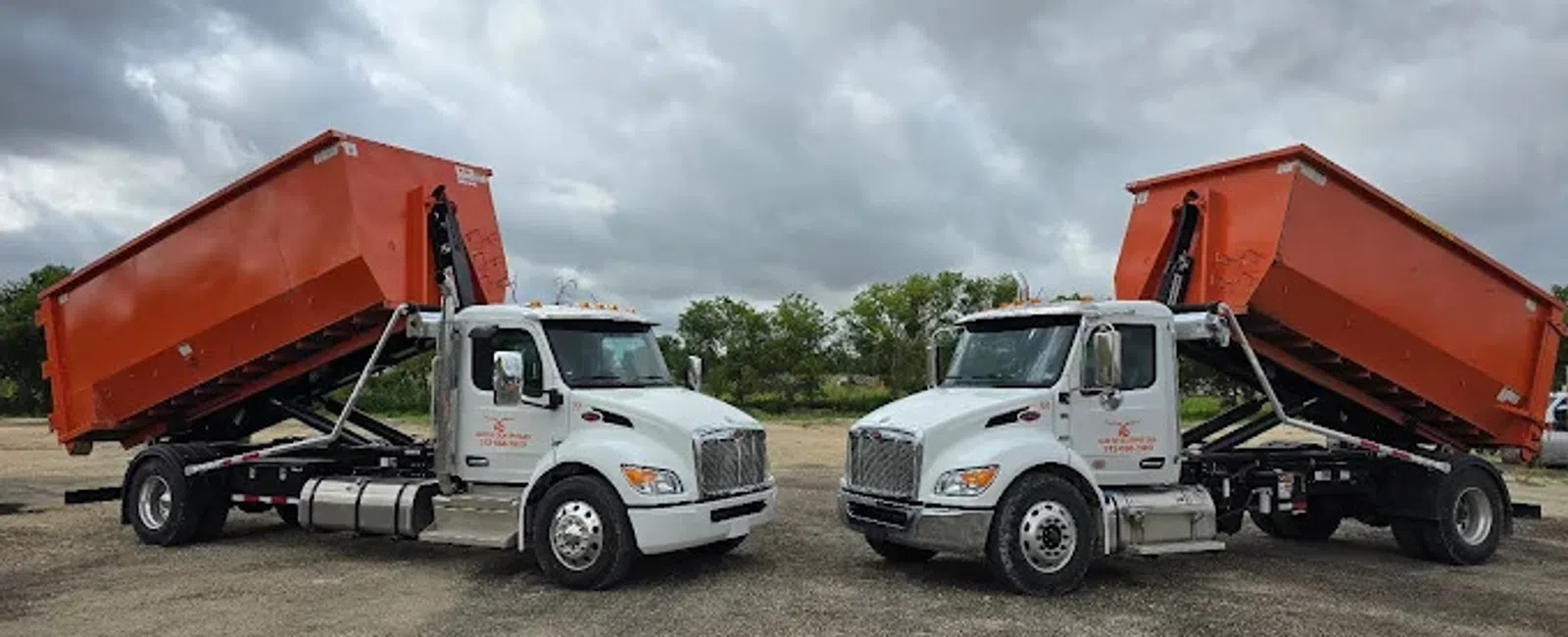 Two white dump trucks with raised orange containers parked on gravel under a cloudy sky with trees in the background