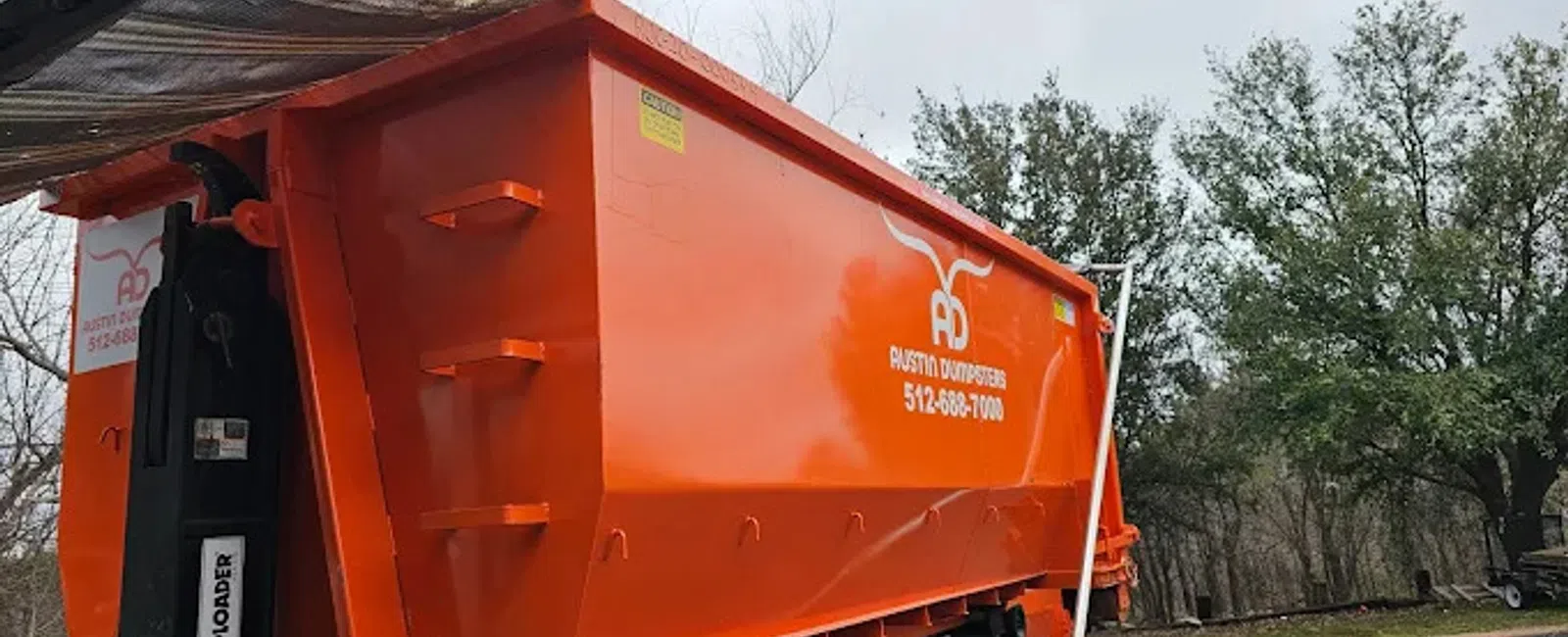 Large orange dumpster trailer with Austin Dumpsters branding parked on a driveway near trees and cloudy sky.