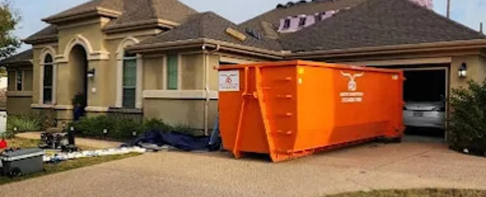 House with roof construction work in progress and large orange dumpster in driveway during daytime.