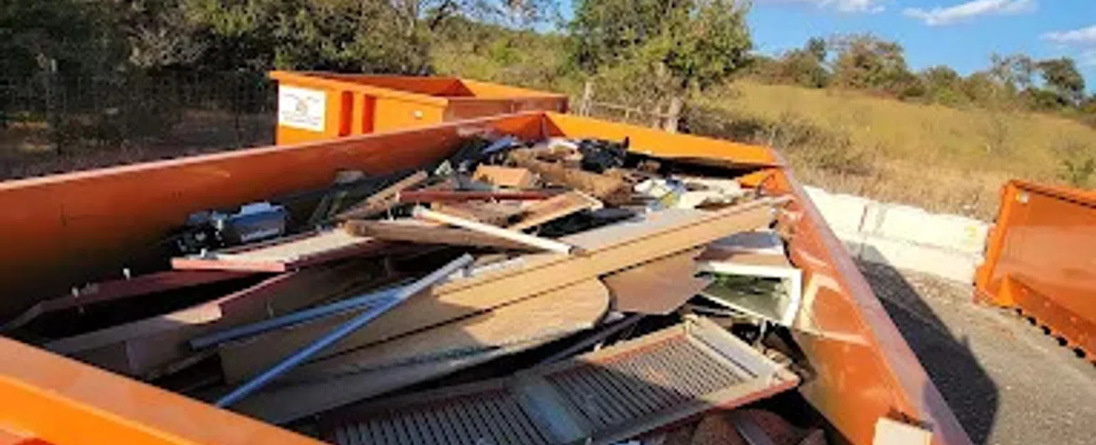 Orange dumpster filled with wooden debris and scrap materials outdoors on a sunny day with trees in background
