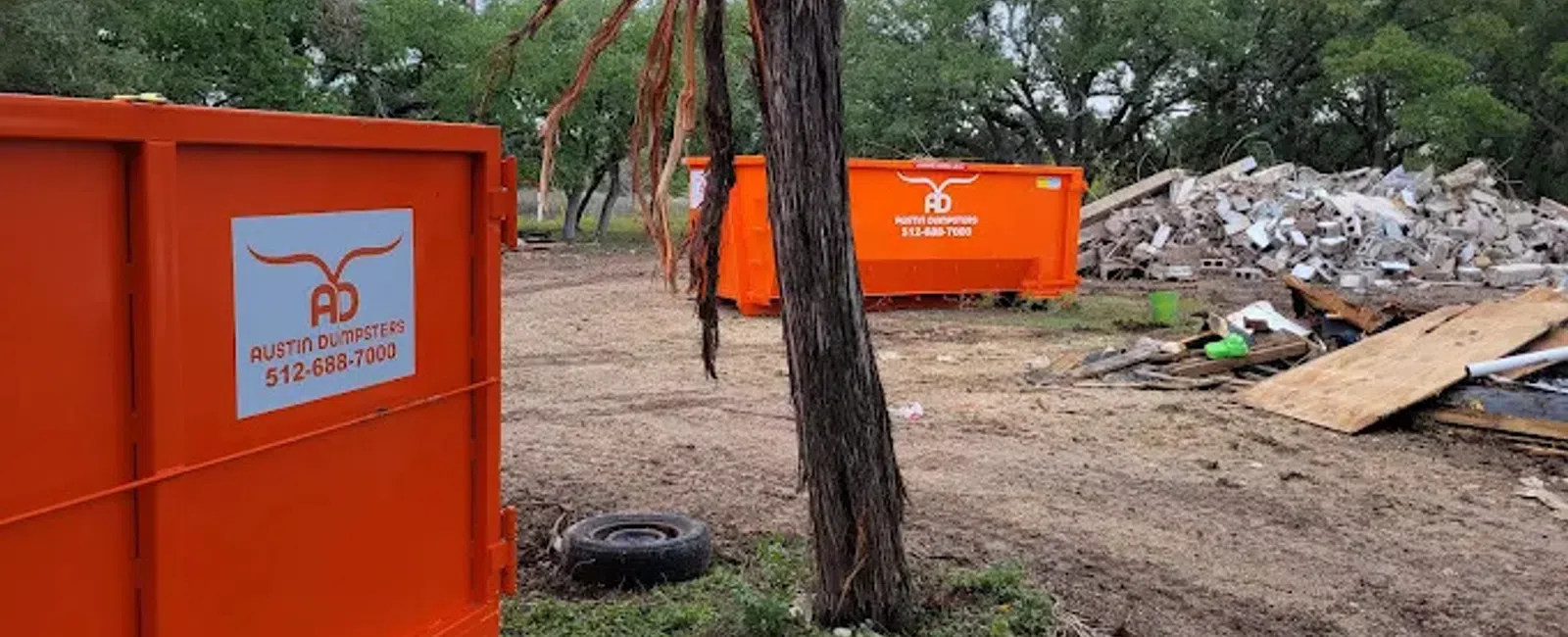 Two bright orange Austin Dumpsters at a debris cleanup site with trees and rubble in the background