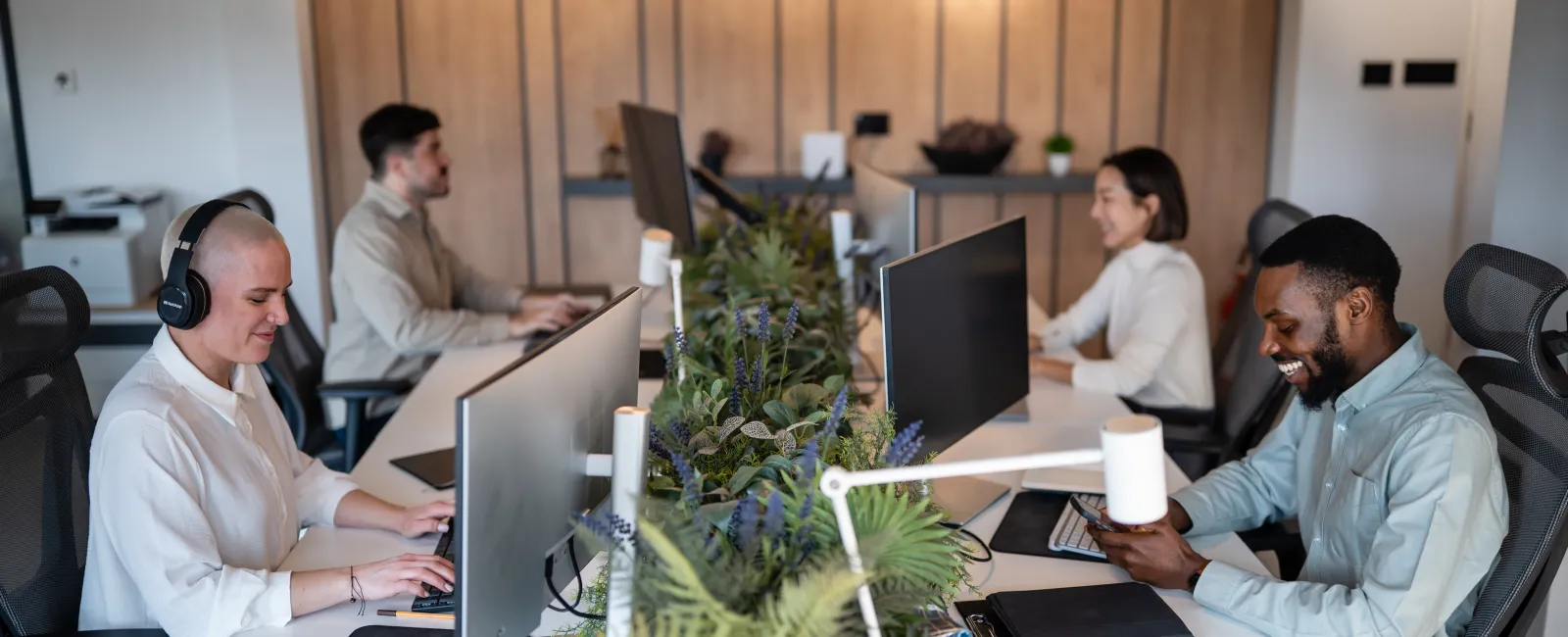 Diverse team working at computers in modern office with desk plants and ergonomic chairs under black pendant lights.