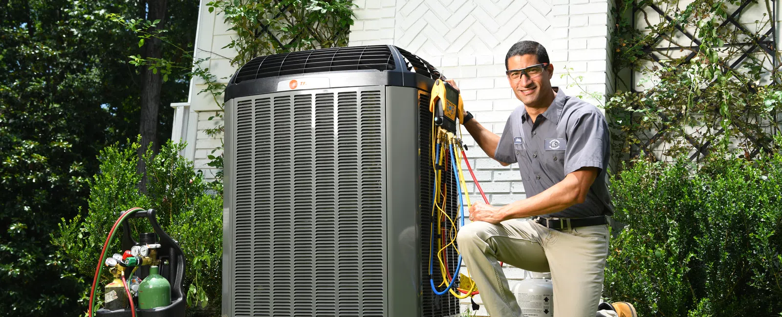 Technician kneeling next to an outdoor HVAC unit performing maintenance with gauges and tools on a sunny day.